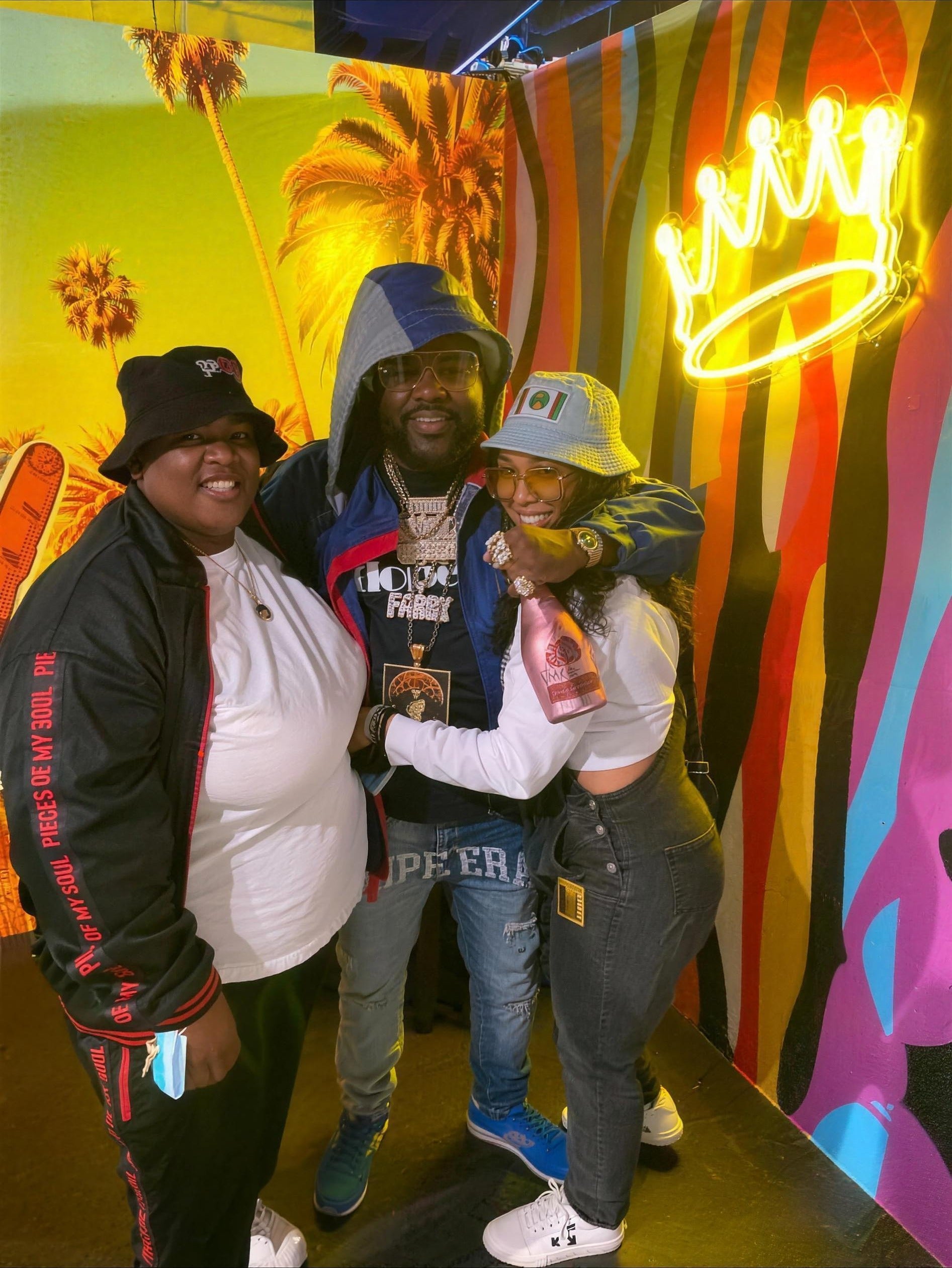 Three friends posing together wearing Pieces of My Soul streetwear in front of a neon crown sign